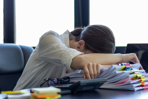 A young secretary is searching through the pile of documents on her desk to prepare for an executive meeting. The secretary looked exhausted from searching through the pile of documents on her desk.