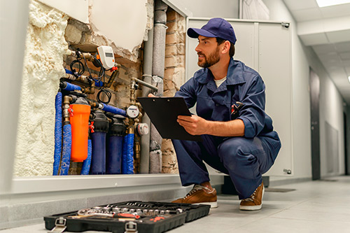 A man in a blue uniform kneels to inspect a water heater, highlighting plumbing issues related to summer water damage in Texas.