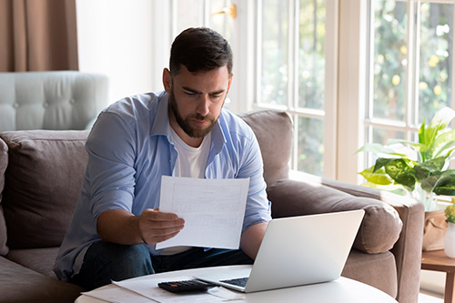 Man on a couch using a laptop, surrounded by papers, analyzing critical insurance settlement mistakes.