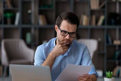 Working with papers. Serious millennial male entrepreneur work at home office alone hold read document hardcopy think on conditions terms. Focused young man engaged in paperwork edit text of agreement