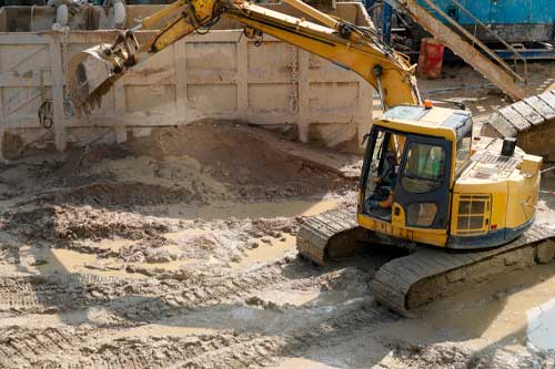 Heavy machinery is busy moving earth at a construction site. The muddy ground shows the impact of the work. Surrounding structures indicate ongoing building efforts.
